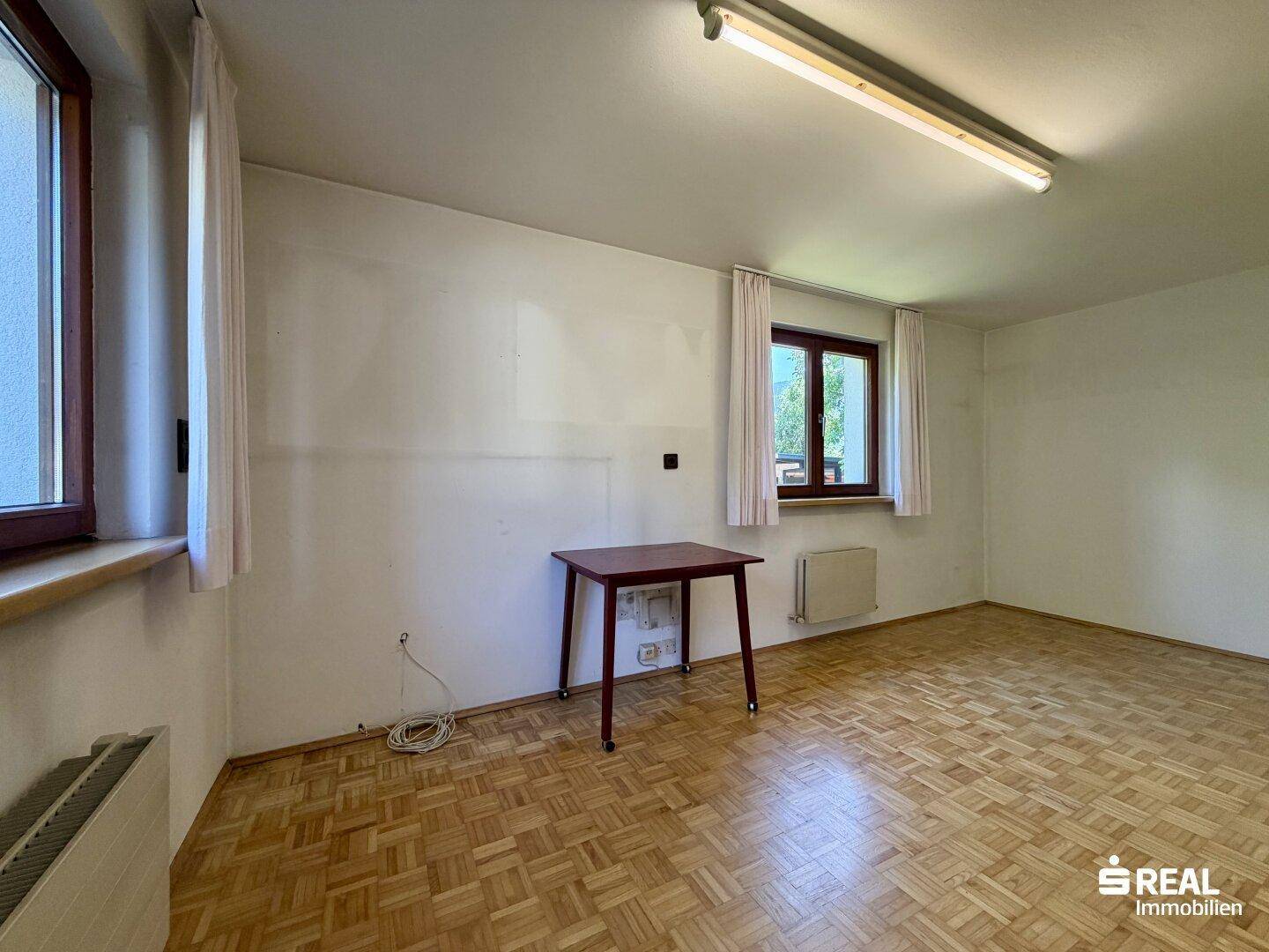 Vielseitiger Raum mit Parkettboden, zwei Fenstern und einfacher Ausstattung.