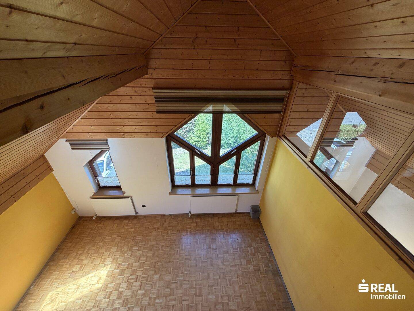 Helles Zimmer im Dachgeschoss mit Holzverkleidung, großen Fenstern und schrägen Wänden.