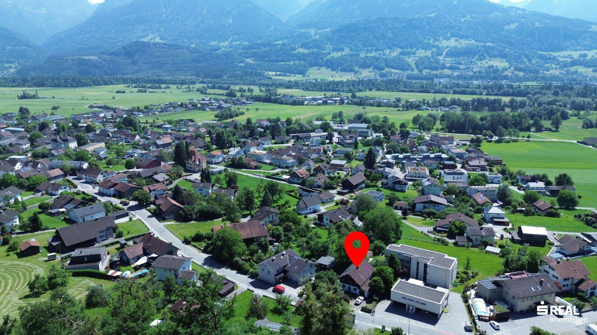 Luftaufnahme einer ländlichen Siedlung mit Bergen im Hintergrund und markiertem Grundstück.