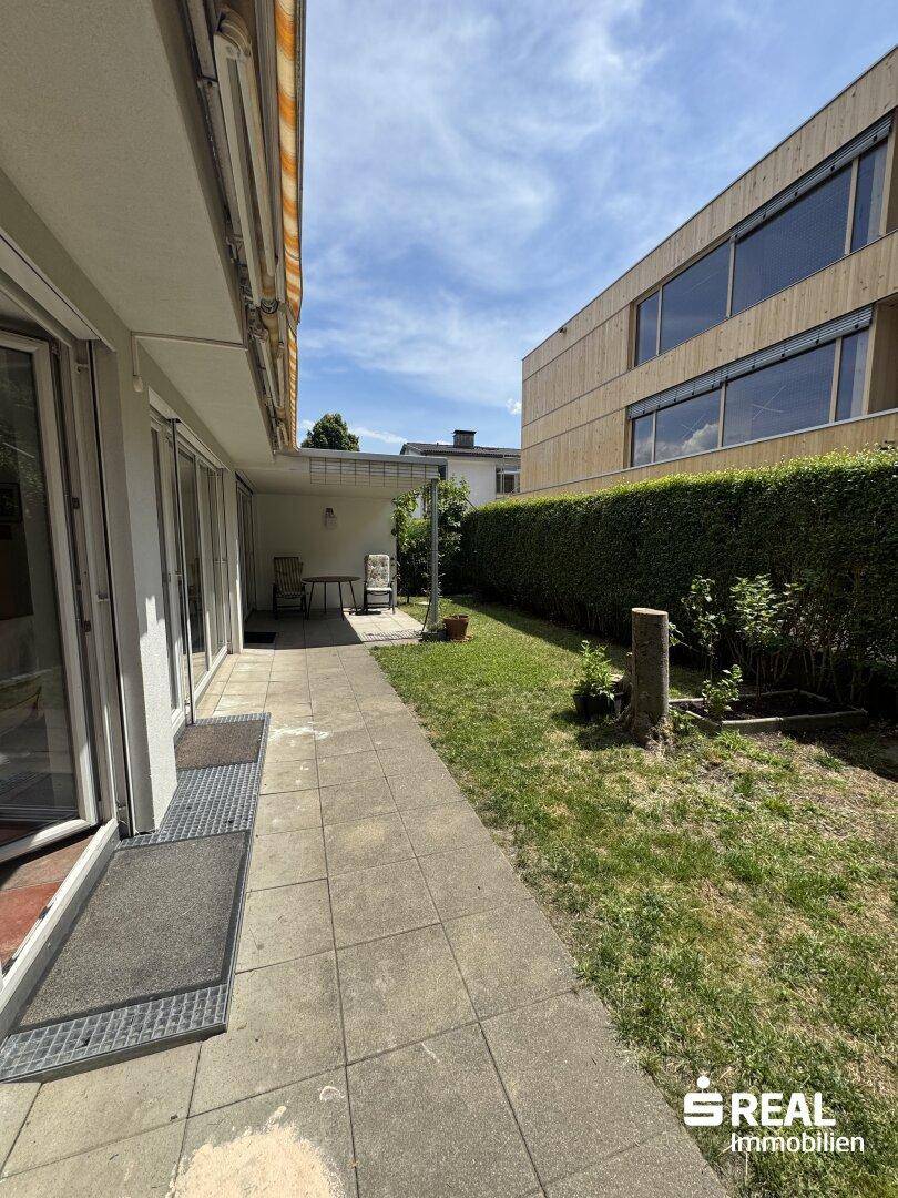 Sonniger Außenbereich mit gepflasterter Terrasse, Rasenfläche und Hecke.