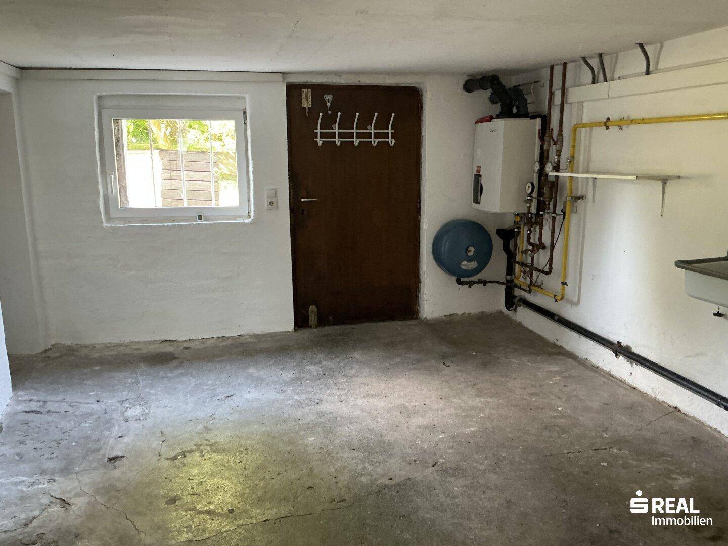 Technikraum im Keller mit Warmwasserbereiter, Gasleitungen und einem kleinen Fenster.