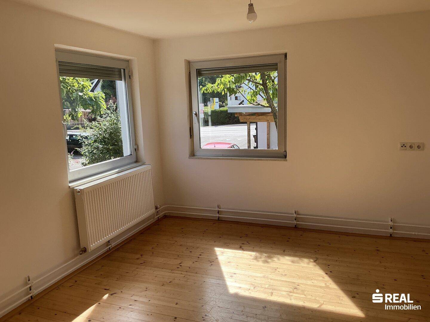 Geräumiges Zimmer mit Holzdielenboden und zwei Fenstern, die viel Tageslicht hereinlassen.