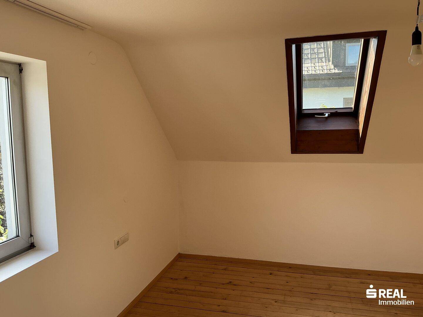 Dachzimmer mit Holzdielenboden, schräger Decke und zwei Fenstern für viel Licht.