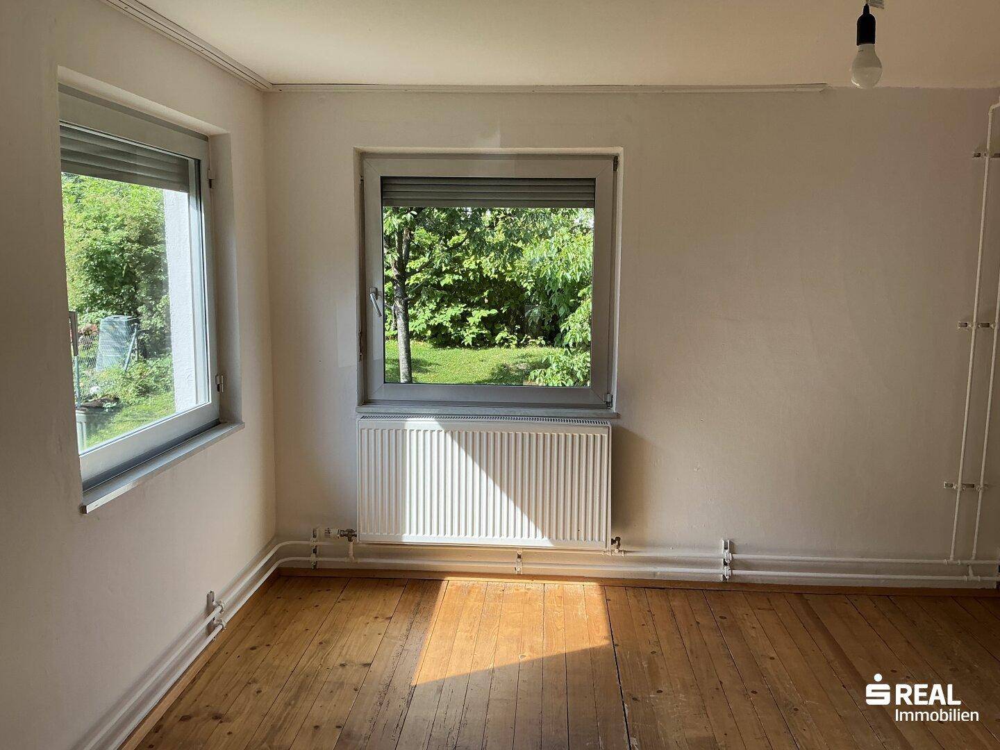 Geräumiges Zimmer mit Holzdielenboden und zwei Fenstern mit Blick ins Grüne.