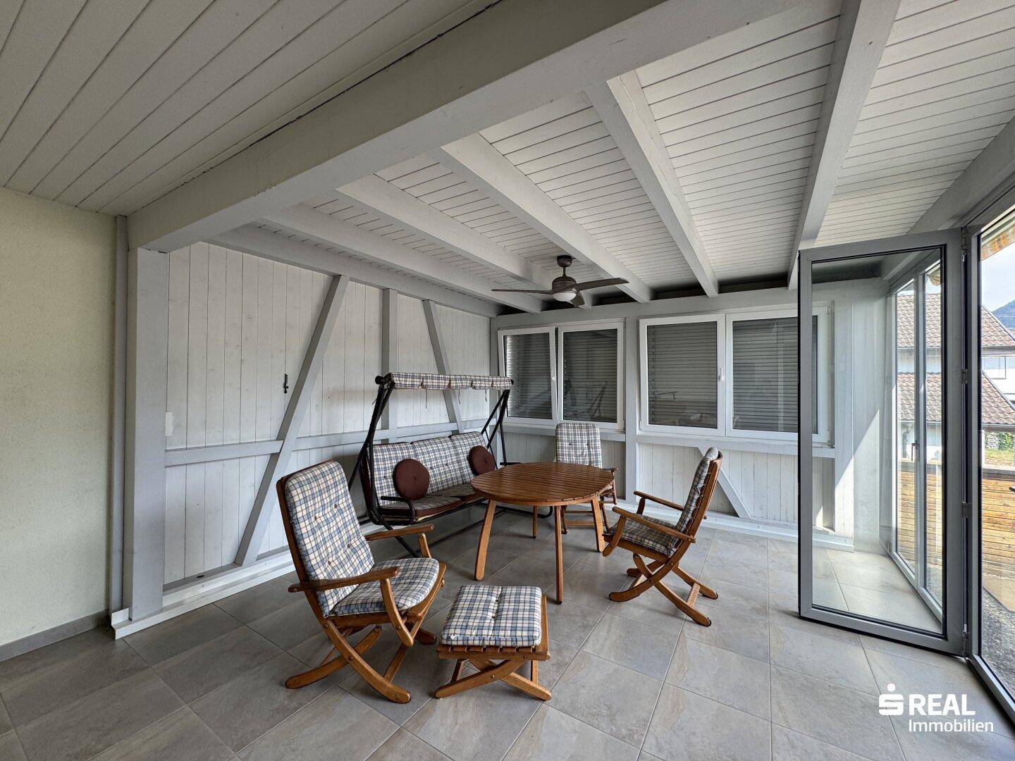 Gemütlicher Wintergarten mit Sitzgelegenheiten, Deckenventilator und Blick auf die Umgebung.