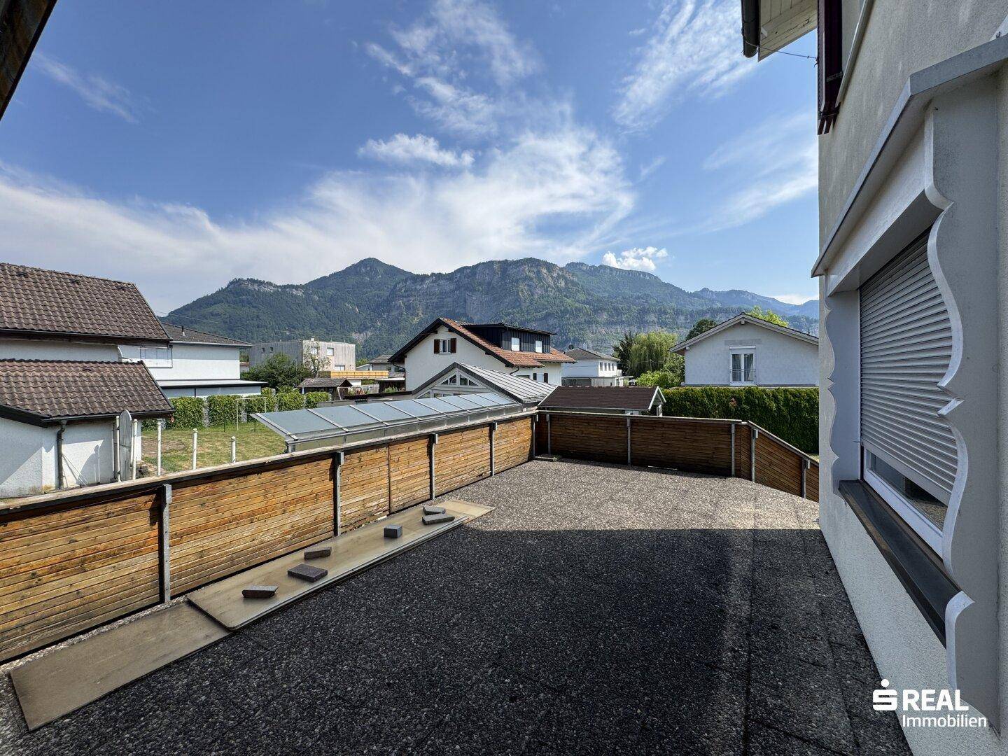 Große gepflasterte Terrasse mit Holzzaun und beeindruckendem Bergblick unter strahlend blauem Himmel.