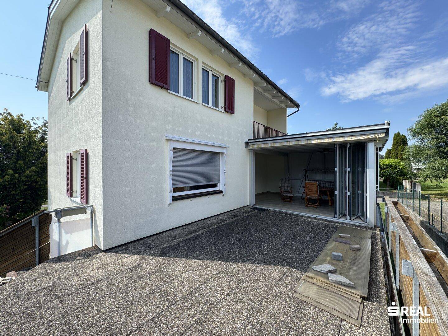Seitenansicht des Hauses mit überdachtem Terrassenbereich und angrenzender gepflasterter Fläche unter blauem Himmel.