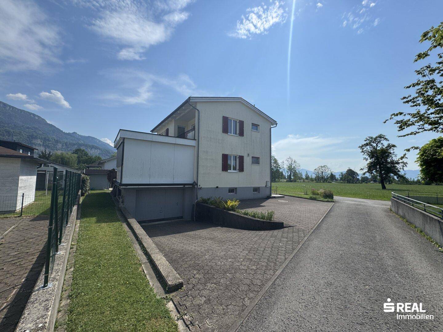 Seitenansicht des Hauses mit gepflasterter Auffahrt und angrenzendem Grünbereich vor ländlicher Kulisse.