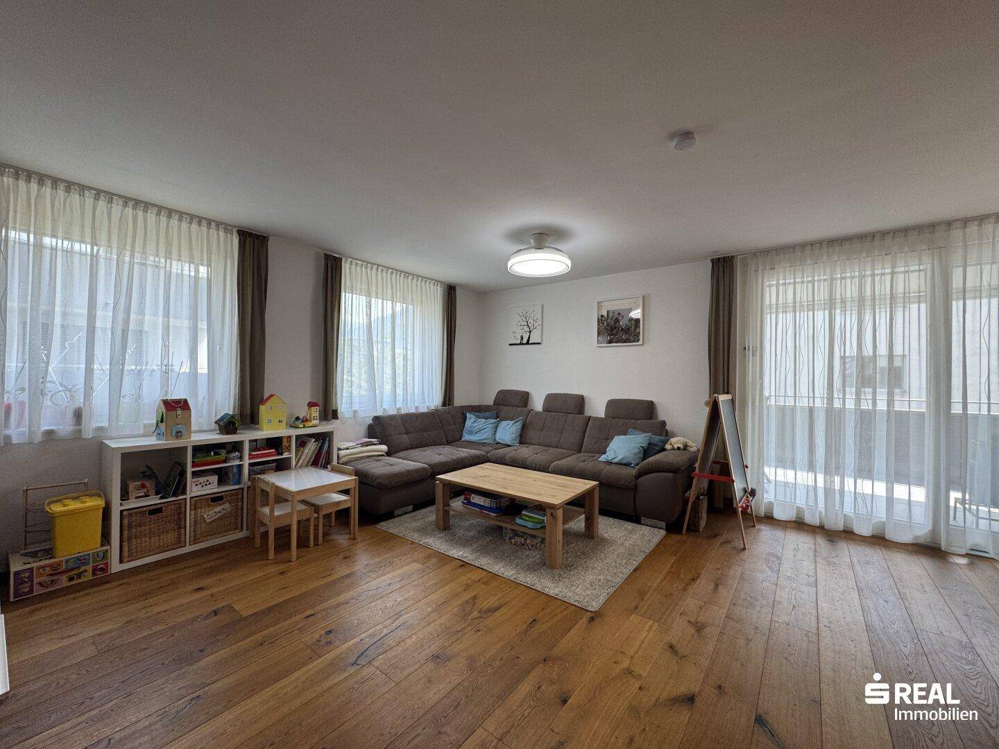 Gemütliches Wohnzimmer mit Holzboden, großem Sofa und viel Tageslicht durch die Fenster.