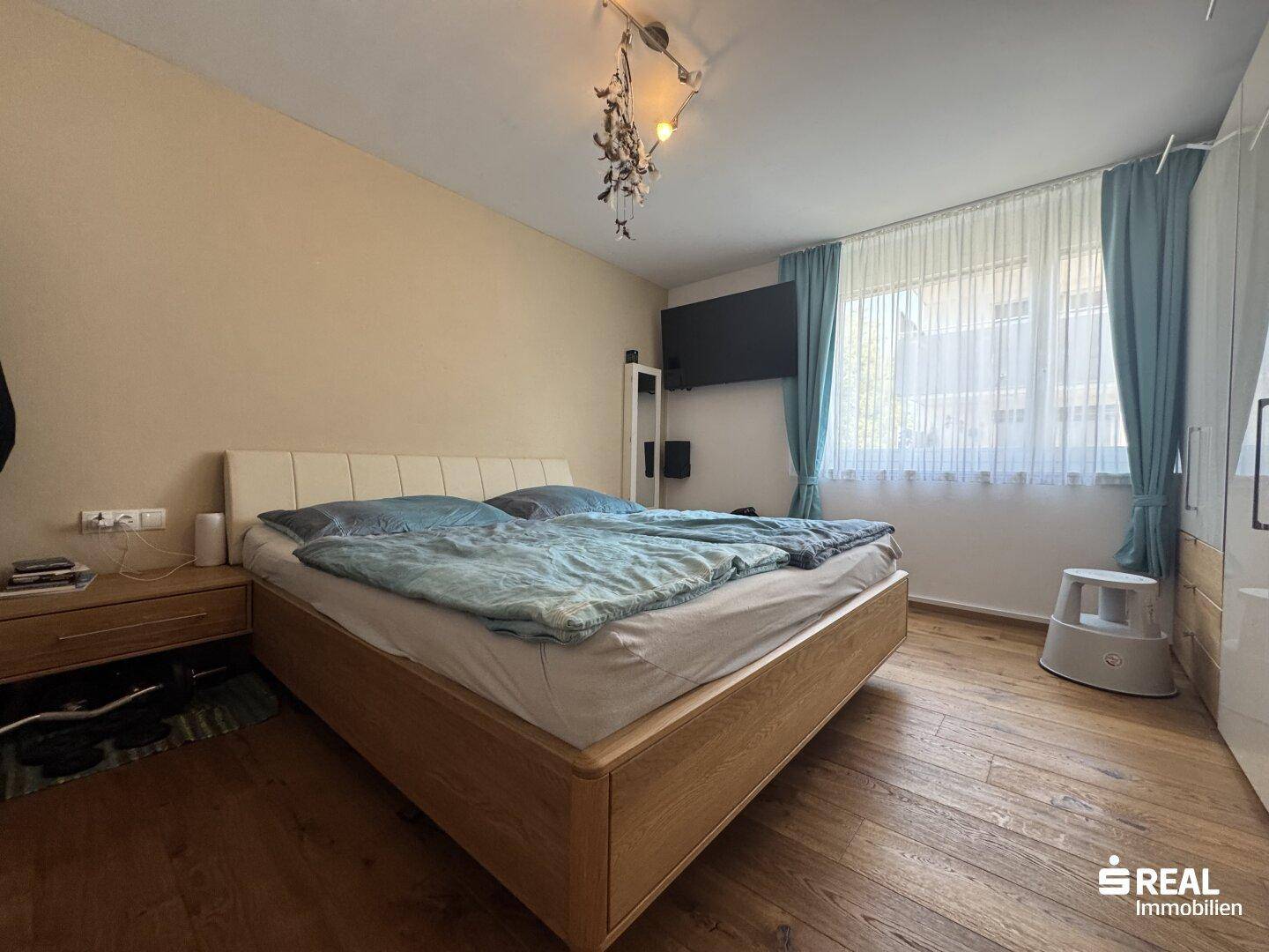 Schlafzimmer mit Doppelbett, Holzboden und großem Fenster, das viel Licht hereinlässt.