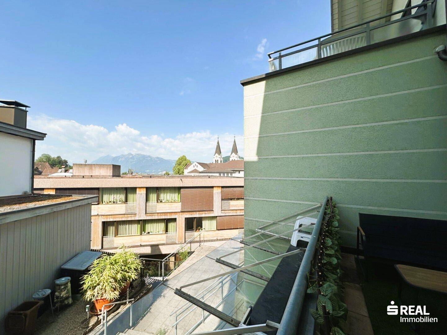 Balkon mit Glasüberdachung bietet Blick auf umliegende Gebäude und die entfernten Berge.