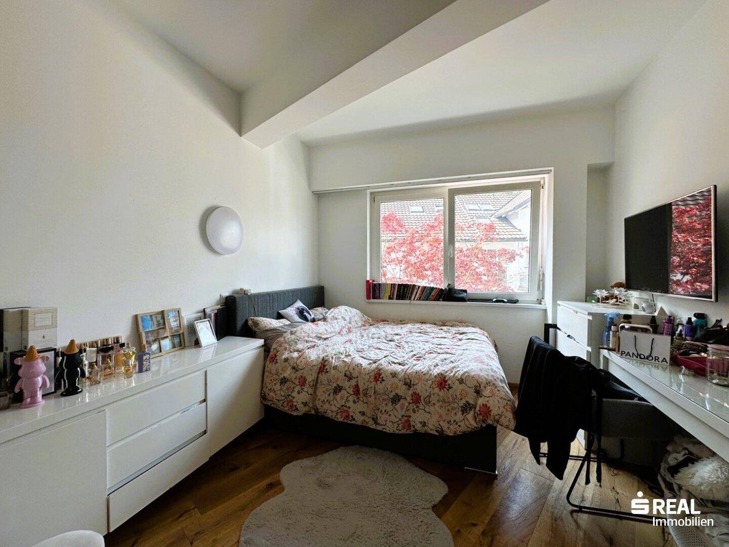 Helles Schlafzimmer mit Holzboden, einem Bett und einem Fenster mit Blick ins Grüne.
