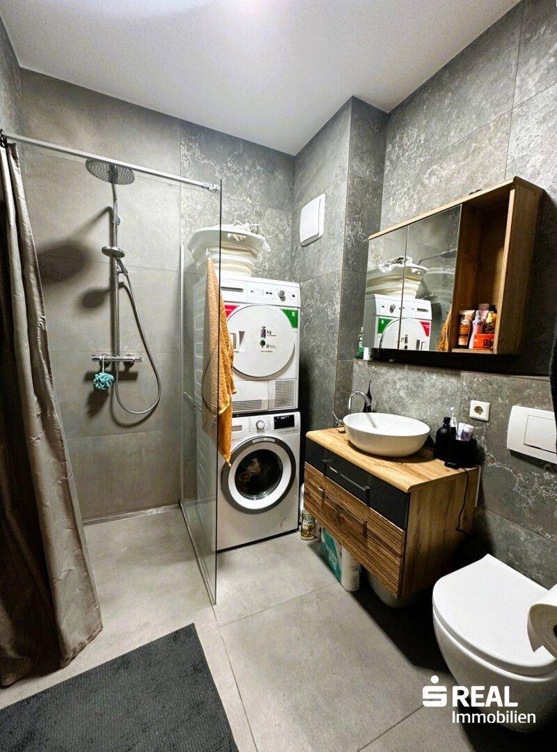Modernes Badezimmer mit grauen Fliesen, Dusche, WC und integrierter Waschmaschine und Trockner.
