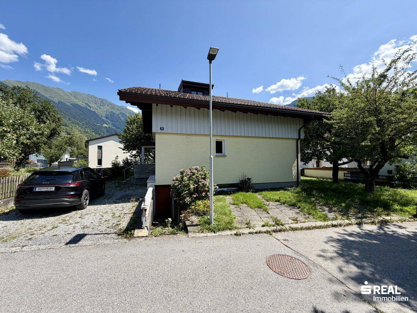 Seitenansicht des Hauses von der Straße mit Parkplatz und Bergblick im Hintergrund.