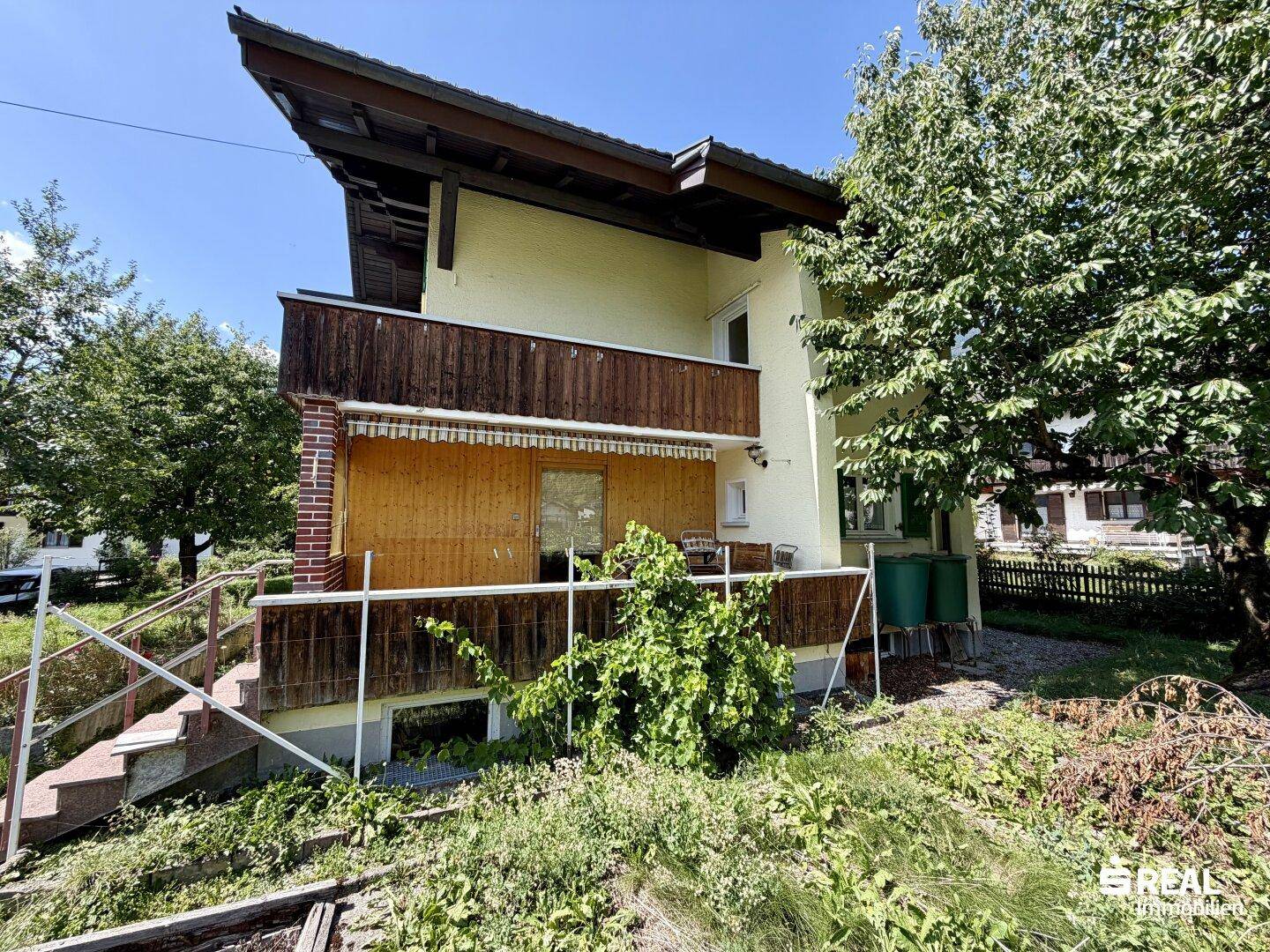 Seitenansicht des Hauses mit Holzbalkon und Treppe, umgeben von viel Grün.