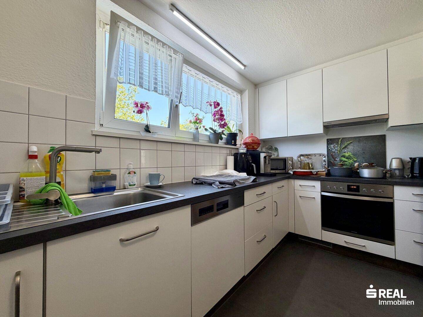 Helle Küche mit weißen Schränken, Spülmaschine und Blick ins Grüne durch das Fenster.