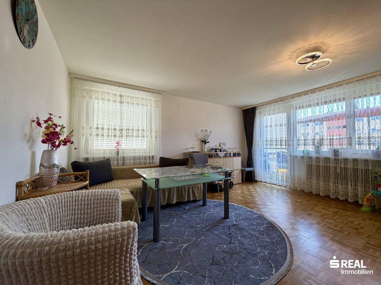 Helles Wohnzimmer mit Parkettboden, Sofa, Couchtisch und großen Fenstern.