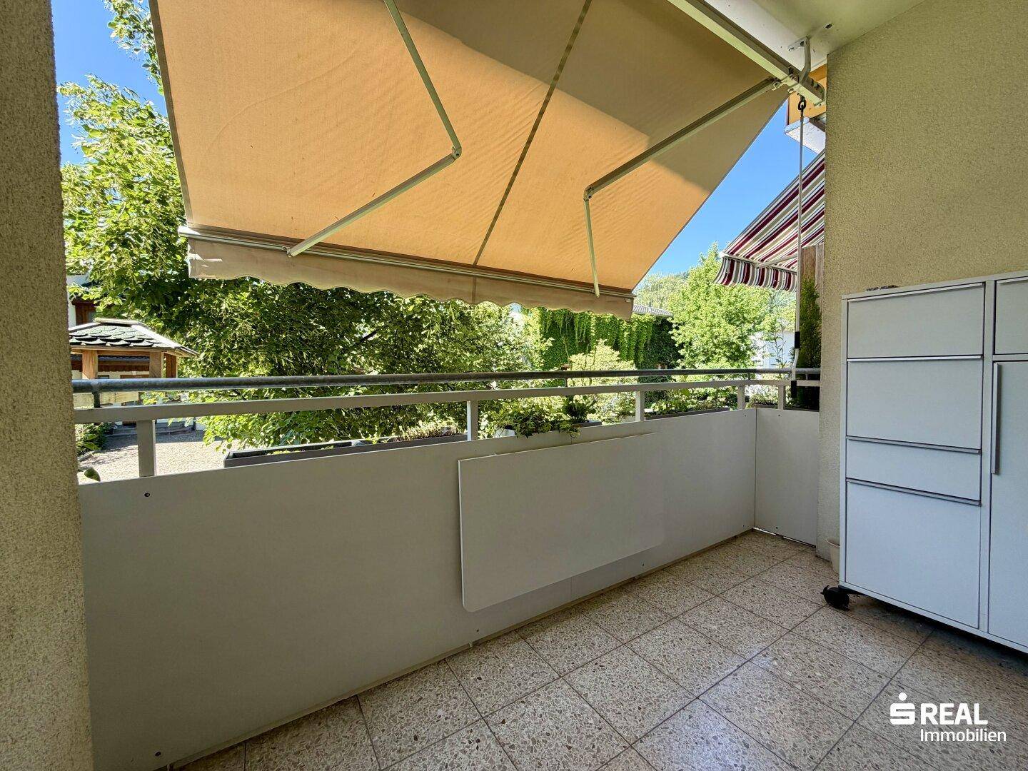 Heller Balkon mit Fliesenboden, Markise und Blick ins Grüne.