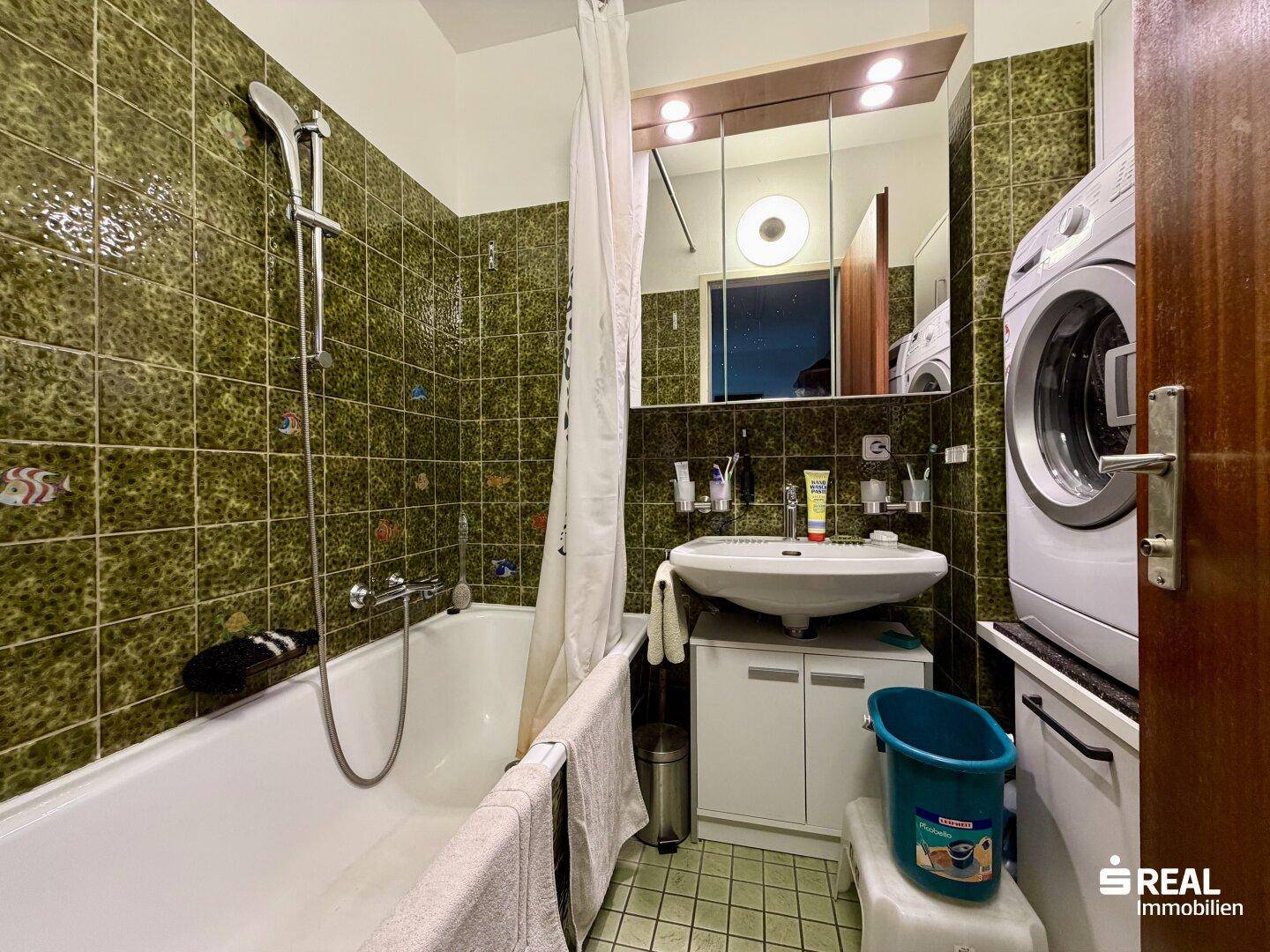Badezimmer mit grünen Fliesen, Badewanne mit Duschfunktion und Waschmaschine.