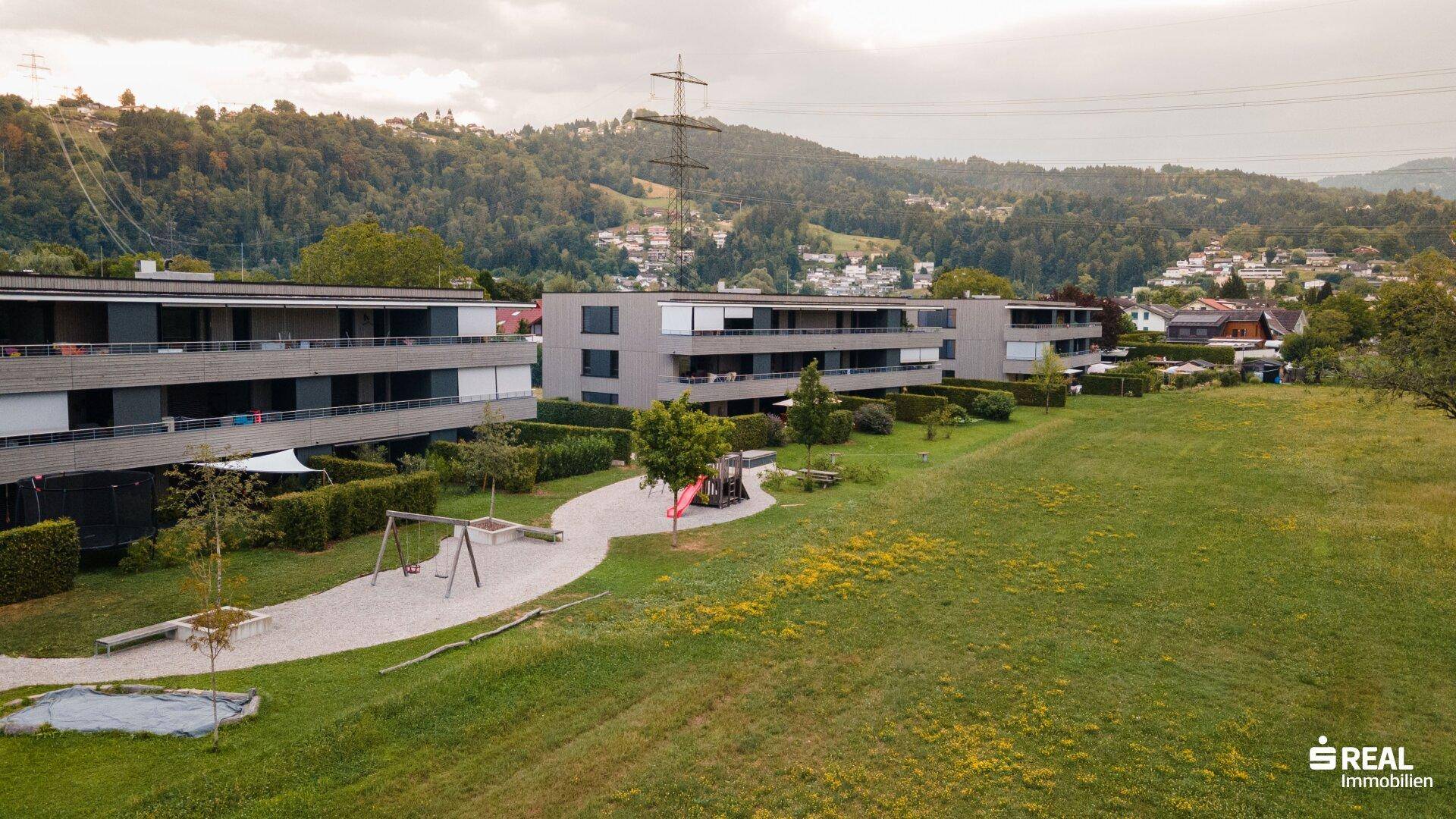 Luftaufnahme einer modernen Wohnanlage mit Spielplatz und weitläufigen Grünflächen in hügeliger Landschaft.