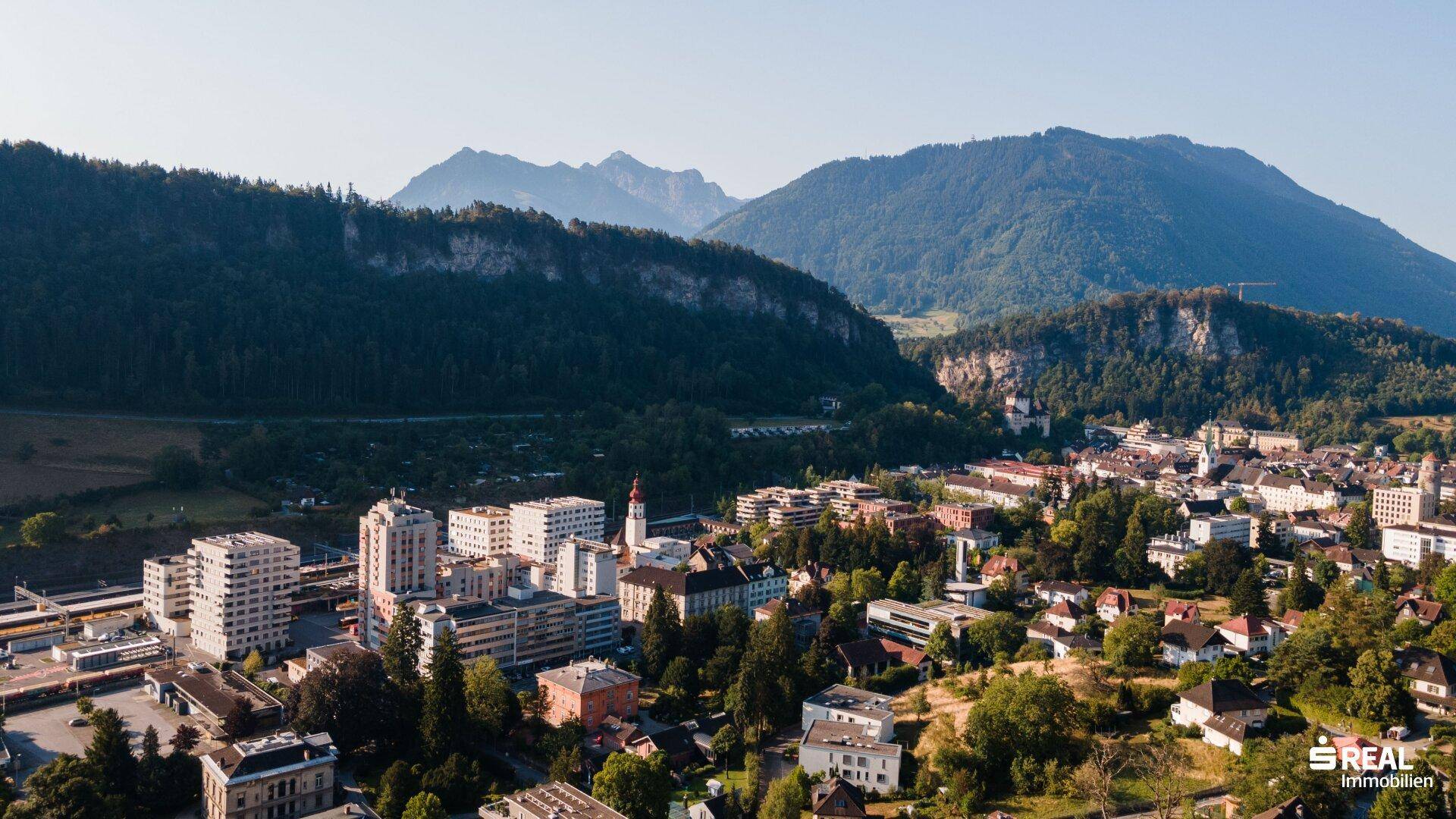 Weitläufiger Blick auf eine Stadt in einem grünen Tal, eingebettet zwischen bewaldeten Bergen und Felsformationen.