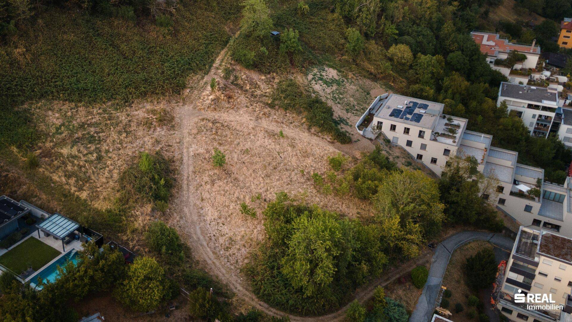 Unbebautes Grundstück an einem Hang, umgeben von modernen Gebäuden mit Flachdächern und einer Zufahrtsstraße.