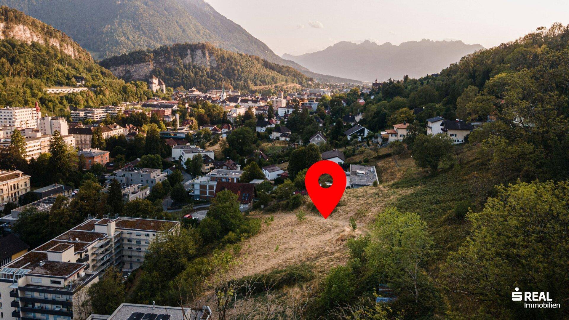 Panoramablick auf eine Stadt in einem Tal, umgeben von bewaldeten Bergen, mit einem roten Standortmarker.