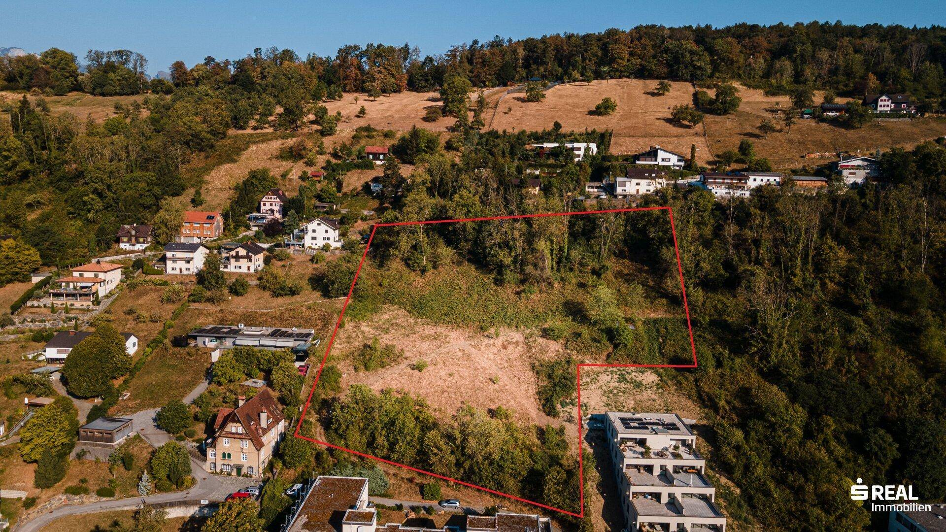 Luftaufnahme eines umgrenzten Grundstücks in einer hügeligen Landschaft mit umliegenden Häusern und viel Grün.