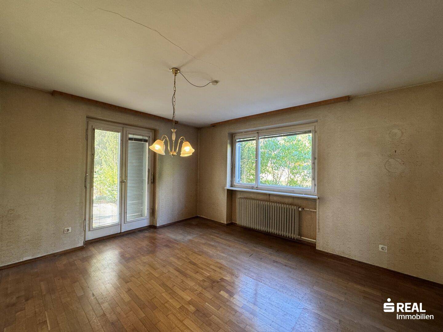 Geräumiges Zimmer mit Parkettboden, Fenster und Zugang zu einem Außenbereich.