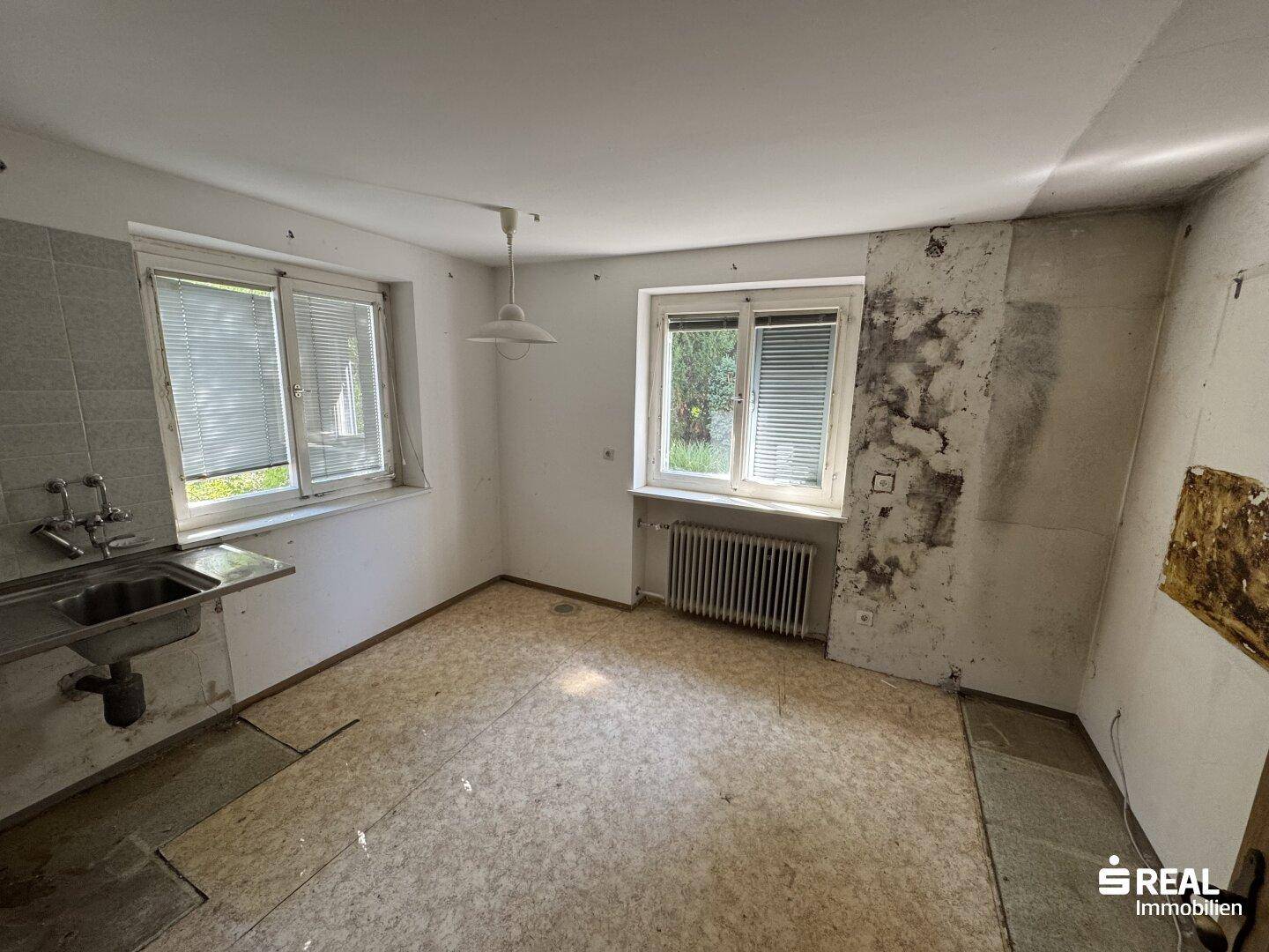 Leere Küche mit Spülbecken, zwei Fenstern und Renovierungsbedarf.