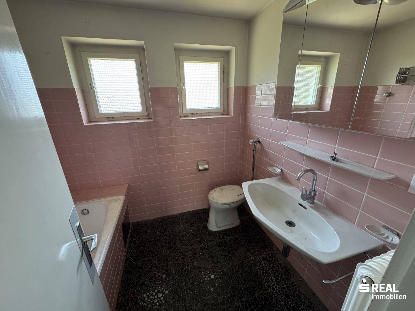 Badezimmer mit rosa Fliesen, Badewanne, Toilette, Waschbecken und zwei Fenstern.
