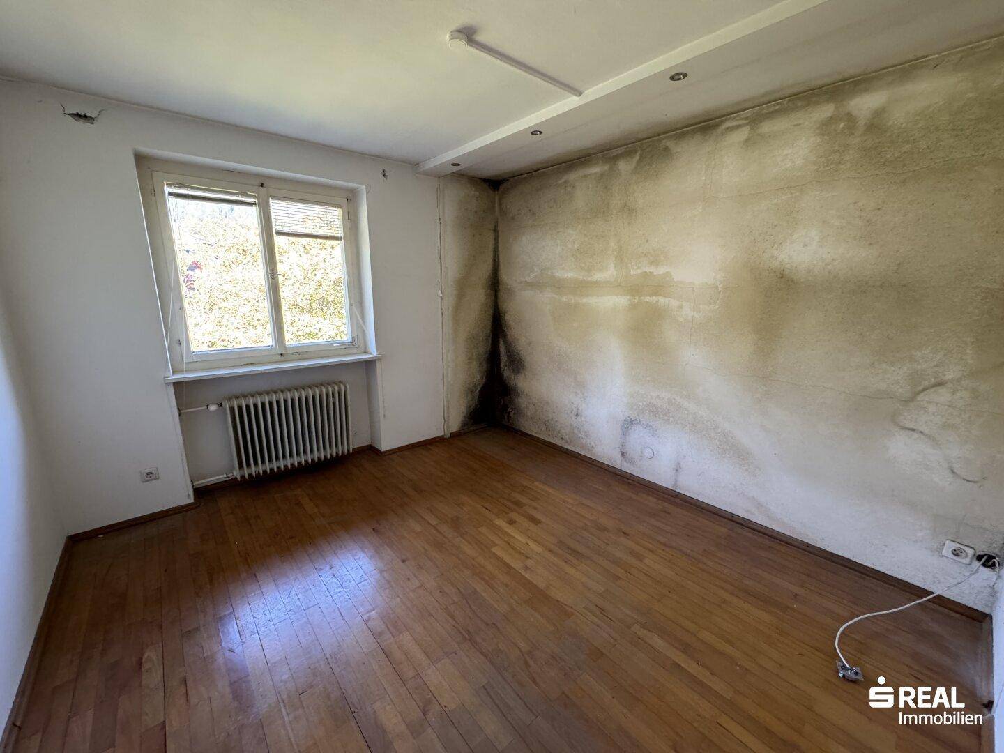 Zimmer mit Parkettboden und Fenster, das Renovierungsbedarf aufgrund von Feuchtigkeit aufweist.