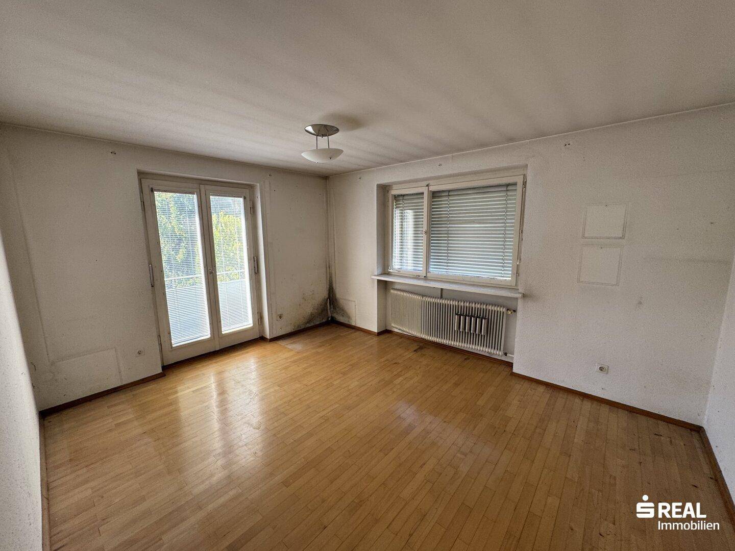 Helles Zimmer mit Parkettboden, großem Fenster und Zugang zu einem Balkon oder einer Terrasse.