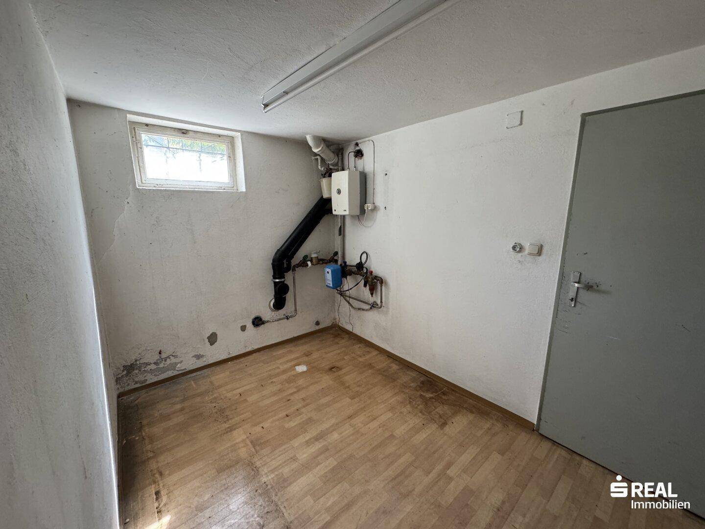 Hauswirtschaftsraum im Keller mit sichtbaren Rohren, Fenster und Holzfußboden.