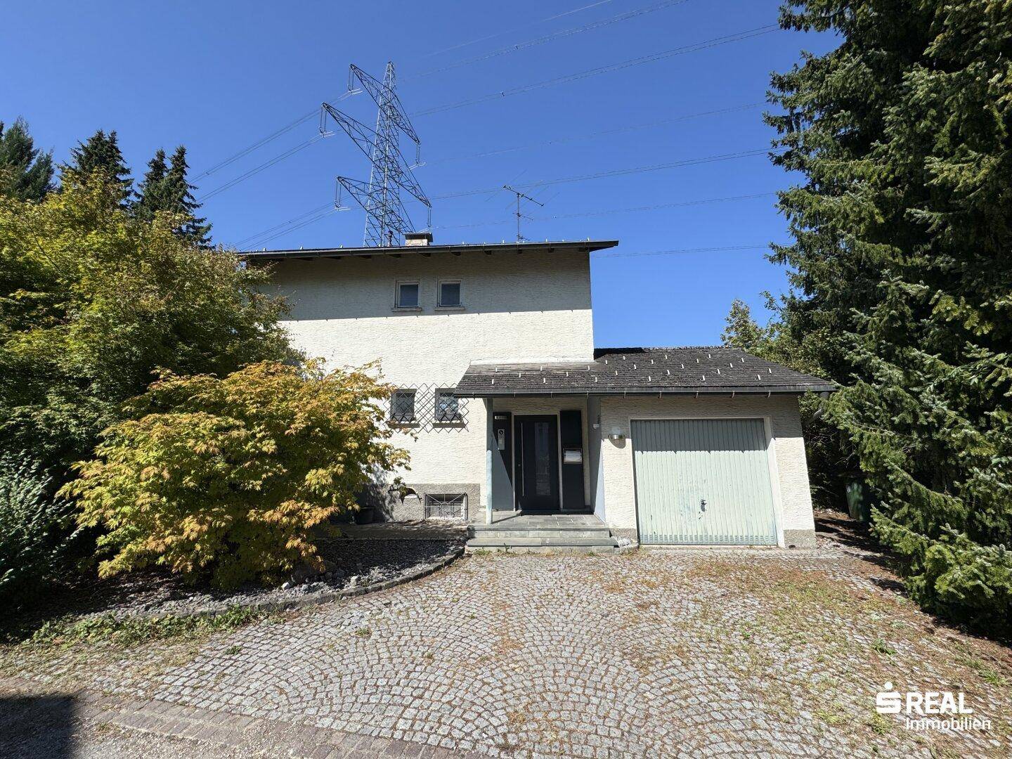 Vorderansicht des Hauses mit integrierter Garage, gepflasterter Auffahrt und Bäumen.