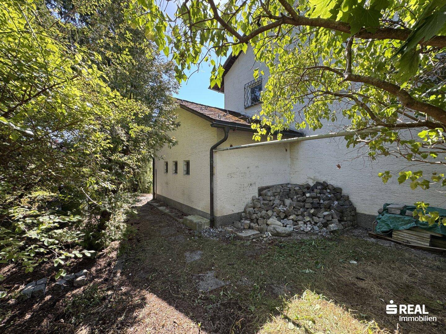 Seitenansicht eines Hauses, umgeben von üppigem Grün und Bäumen, mit einem Steinhaufen an der Wand.