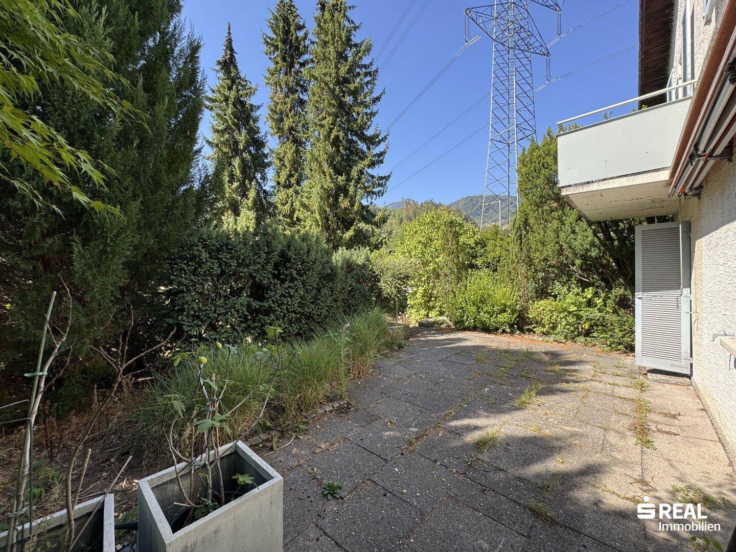 Große gepflasterte Terrasse umgeben von Bäumen und Sträuchern, mit Blick auf die Landschaft und einen Strommast.