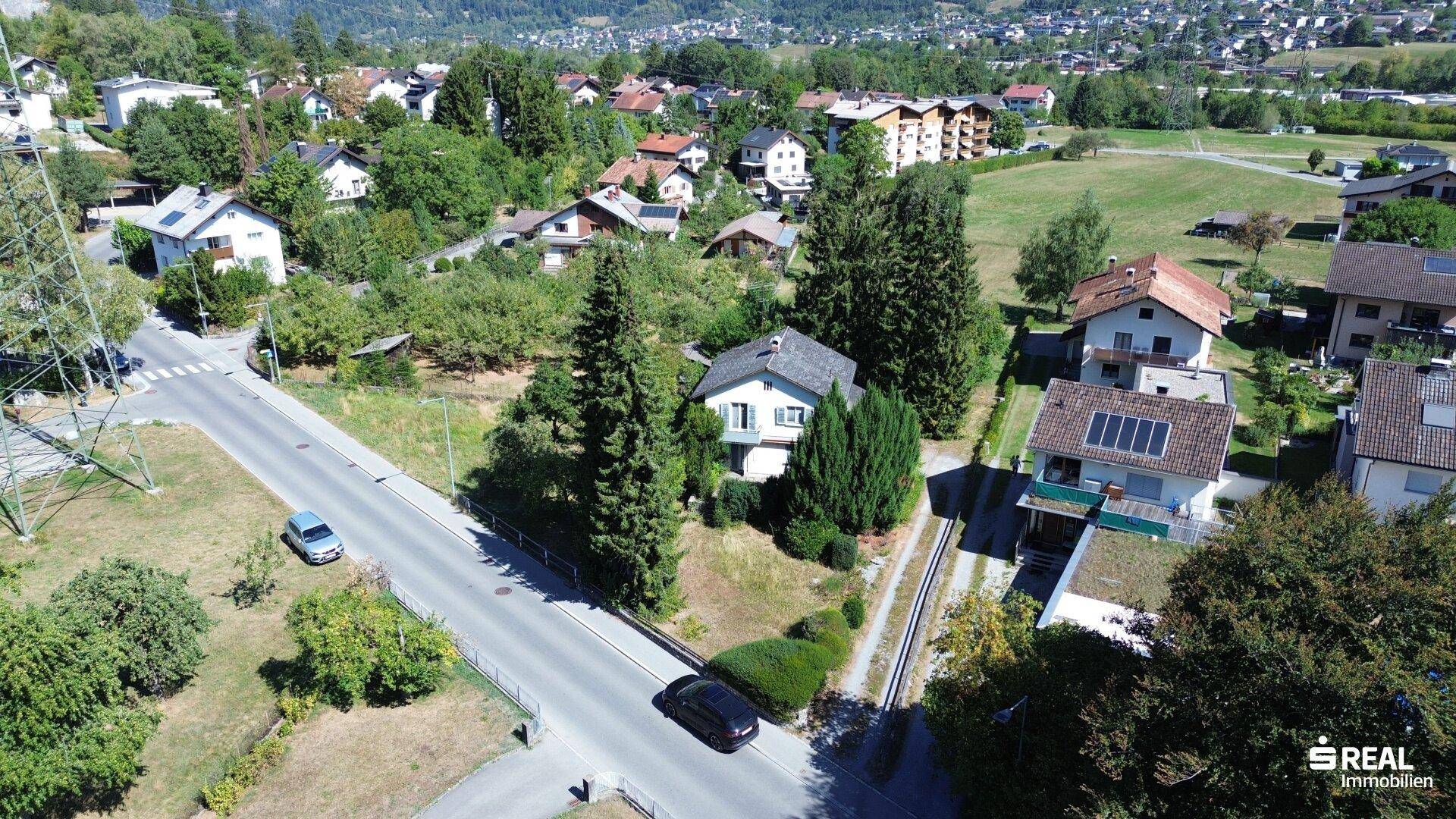 Luftaufnahme einer Wohnsiedlung mit Häusern, Straßen und viel Grün, umgeben von Bergen.