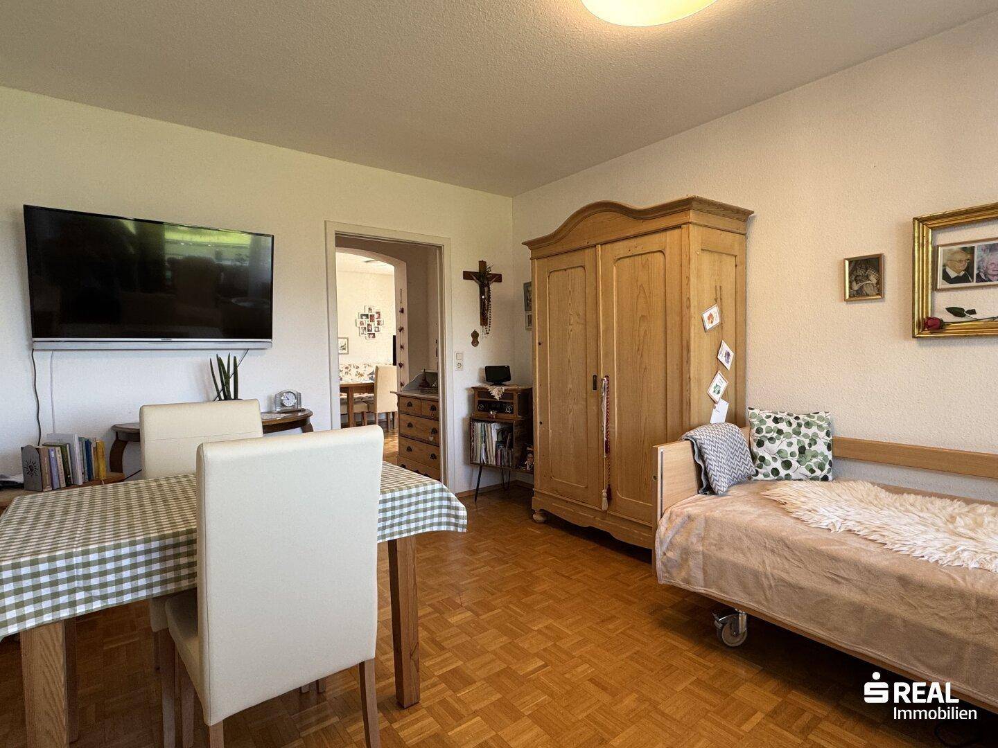 Wohnbereich mit Holzboden, Fernseher und traditionellem Holzschrank, offen zum Essbereich.