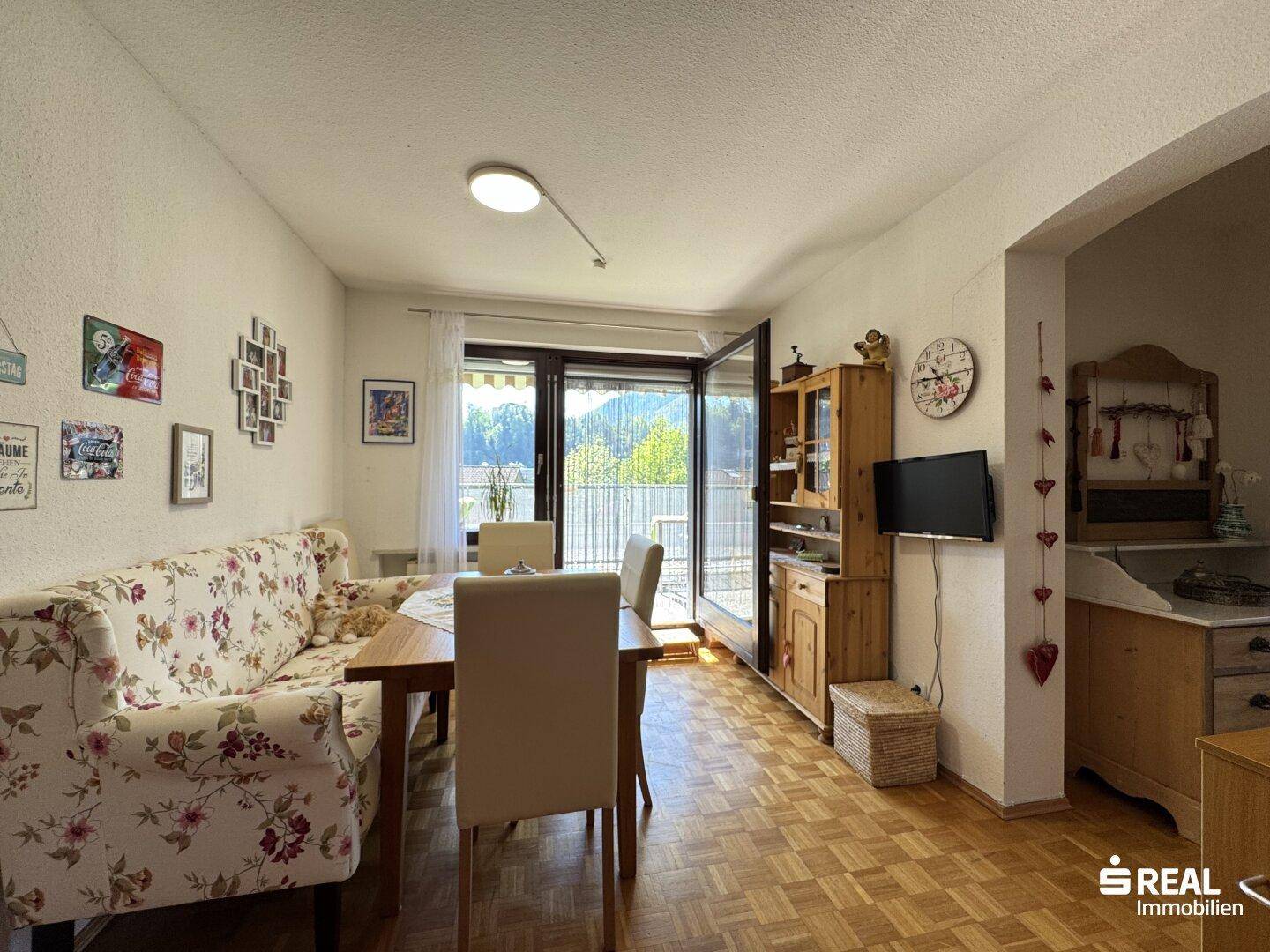 Gemütlicher Wohn-Essbereich mit floralem Sofa, Holztisch und Balkonzugang.