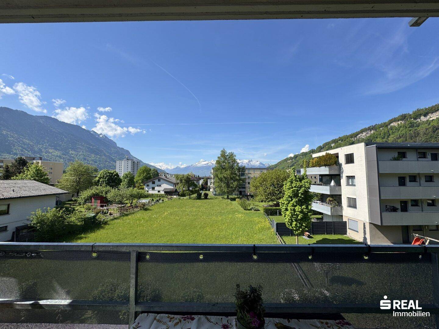 Blick vom Balkon auf eine grüne Wiese mit Bergen im Hintergrund unter blauem Himmel.