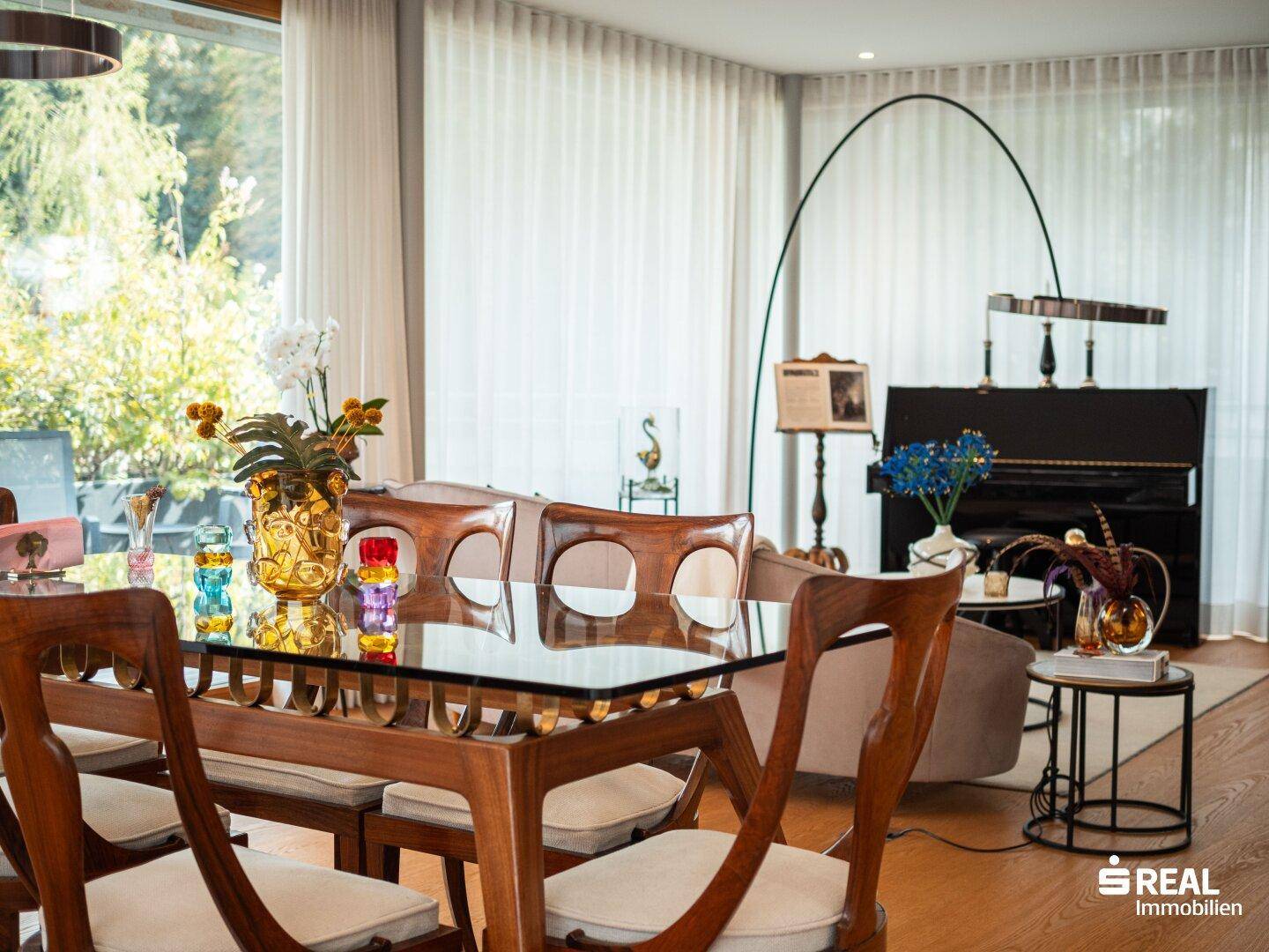 Essbereich mit Holztisch, Glasplatte und Stühlen, umgeben von großen Fenstern.