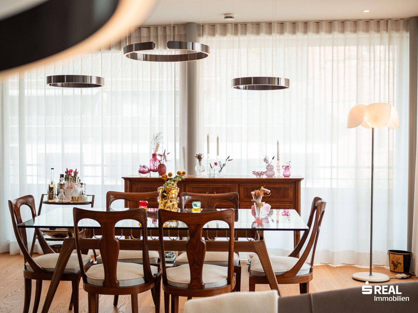 Essbereich mit Holztisch und Stühlen, umgeben von großen Fenstern und moderner Beleuchtung.