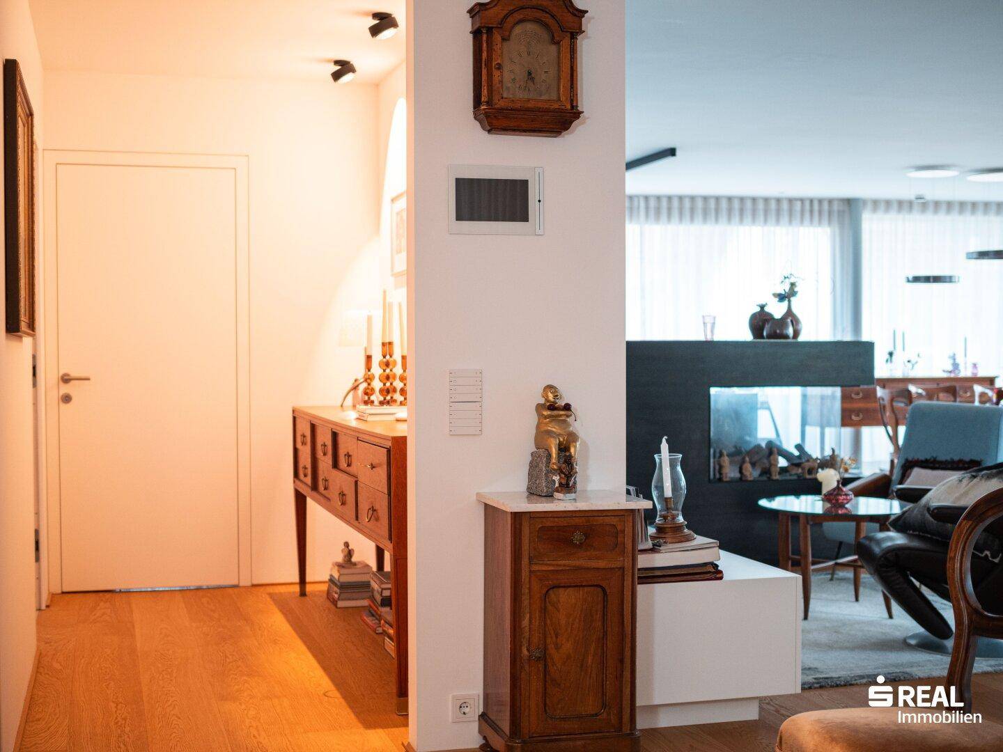 Heller Flur mit Holzboden, antiker Kommode und Blick auf den modernen Wohnbereich mit Kamin.