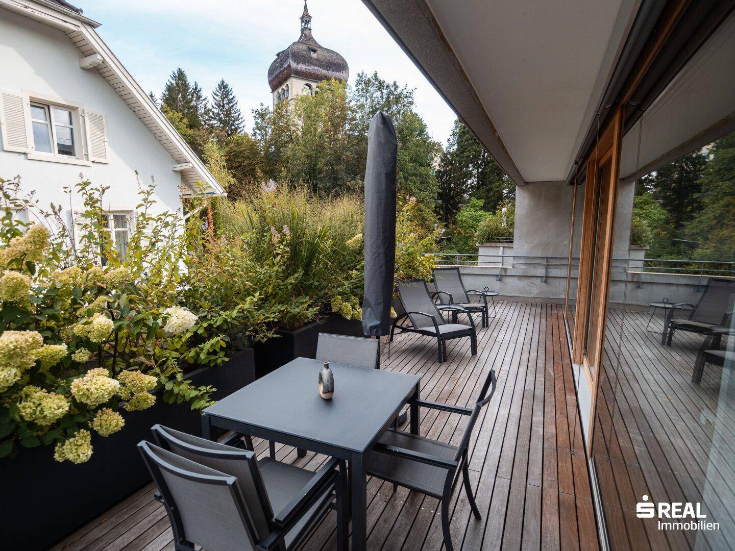 Große Holzterrasse mit Essbereich und Liegestühlen, umgeben von Pflanzen und mit Blick auf einen Kirchturm.