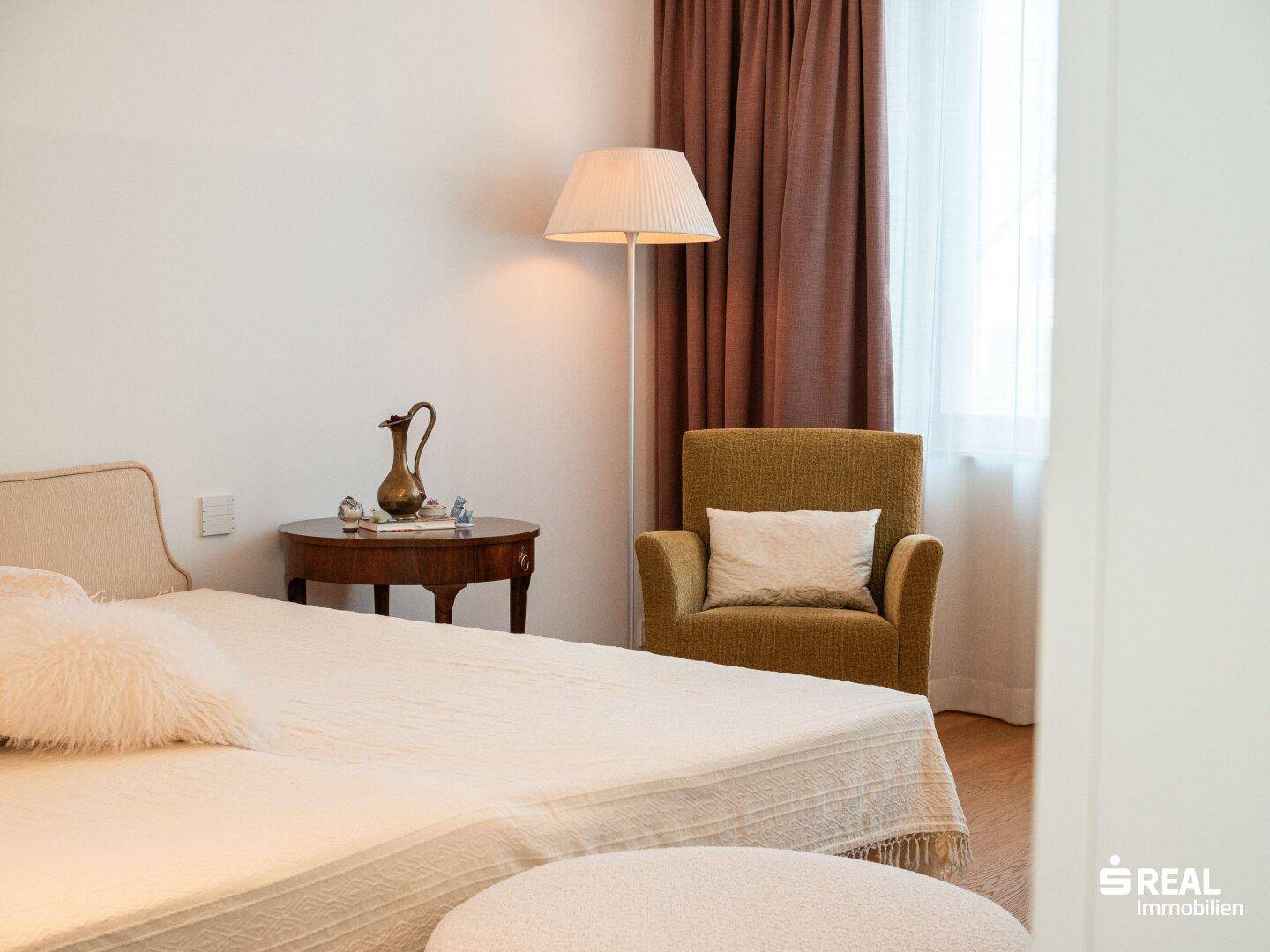 Gemütliches Schlafzimmer mit beigem Bett, grünem Sessel und braunen Vorhängen, hell und einladend gestaltet.