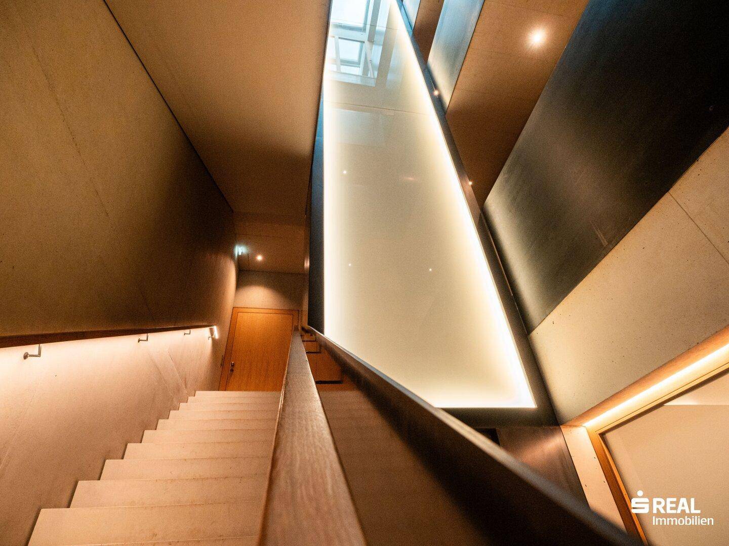 Modernes Treppenhaus mit Betonwänden und einer beeindruckenden, beleuchteten Glaswand, die für Helligkeit sorgt.