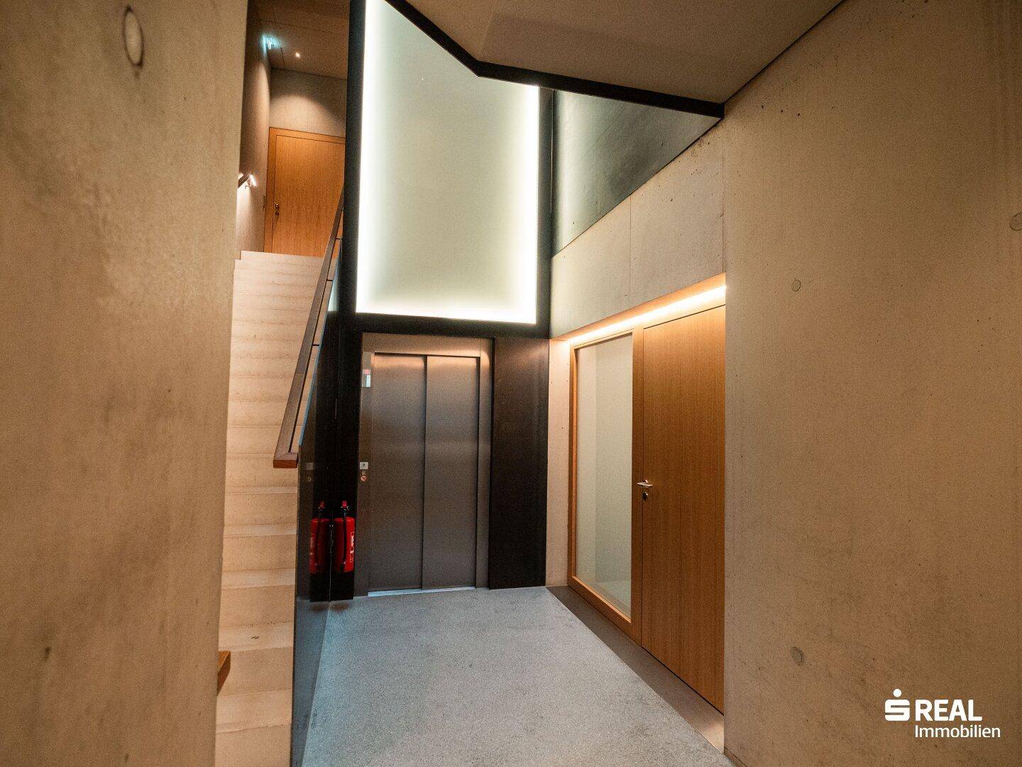 Heller Eingangsbereich mit modernem Aufzug, Treppe und Betonwänden, die ein zeitgemäßes Design unterstreichen.