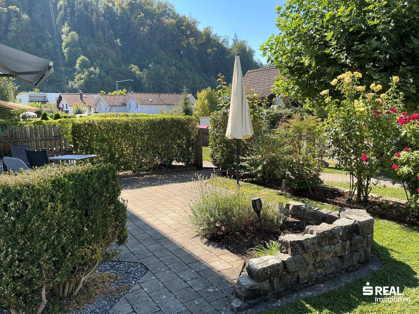 Sonniger Terrassenbereich mit Gartenmöbeln, umgeben von Hecken und Blumenbeeten.