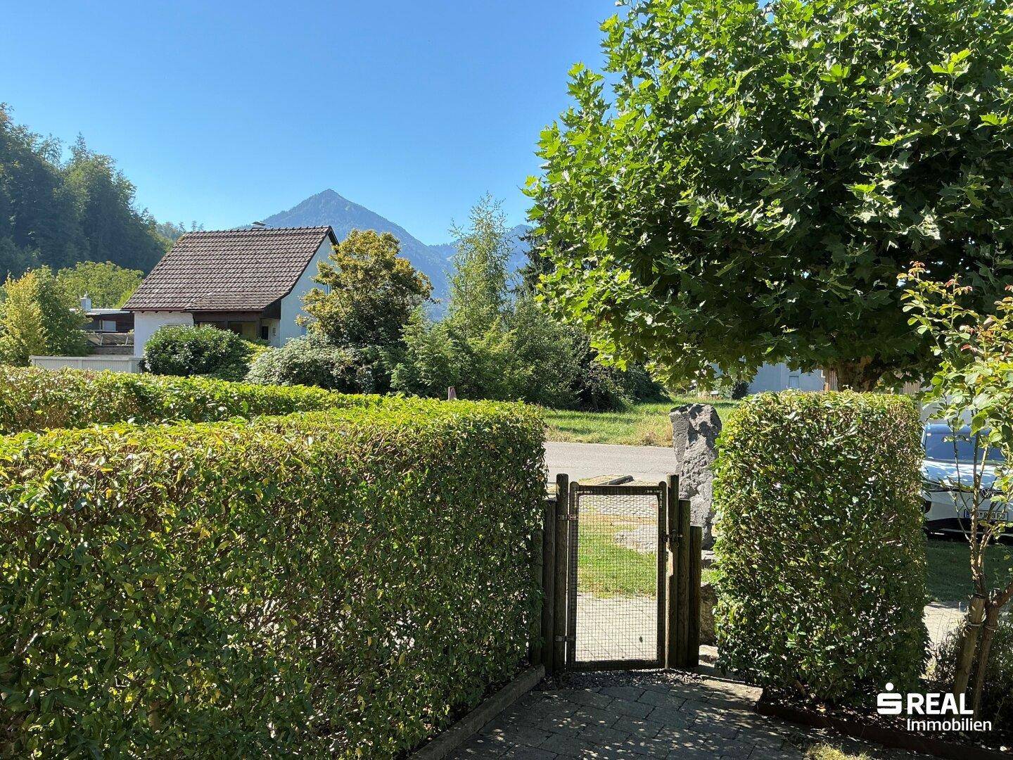 Blick aus dem Garten über eine Hecke hinweg auf die umliegende Berglandschaft.